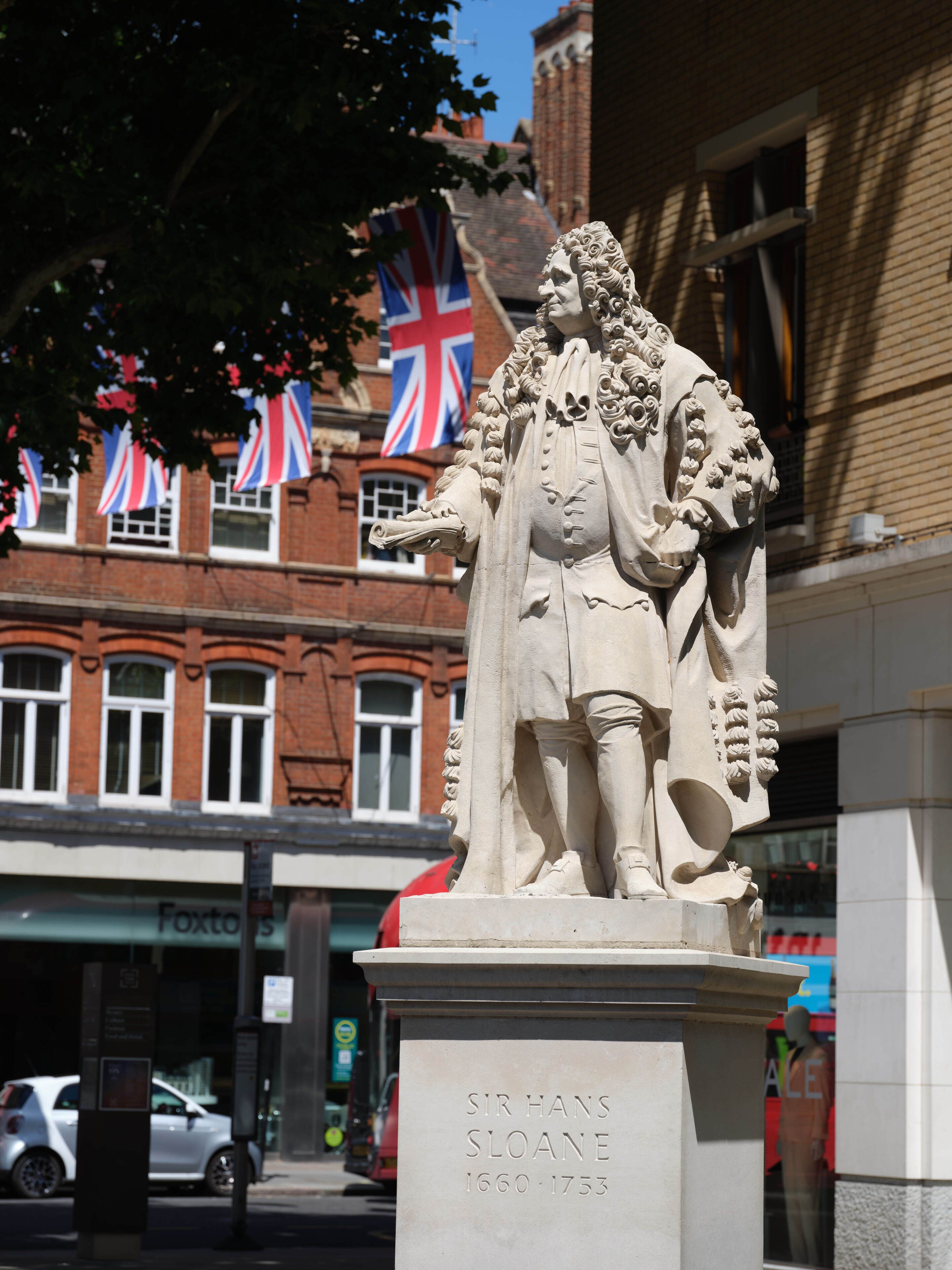 Una estatua de Sir Hans Sloane con una placa que reza 'Sir Hans Sloane 1660-1753' frente a un edificio con banderas británicas.