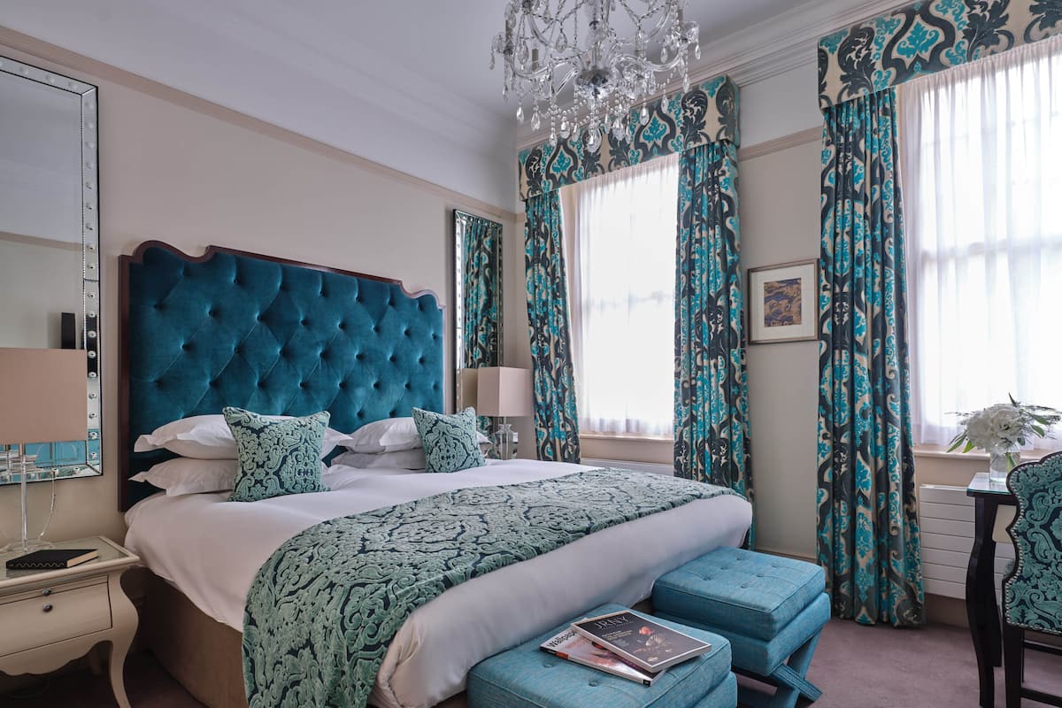 A bedroom with a tufted headboard, a large bed with patterned bedding, a nightstand, a dresser, a chair, and a chandelier.