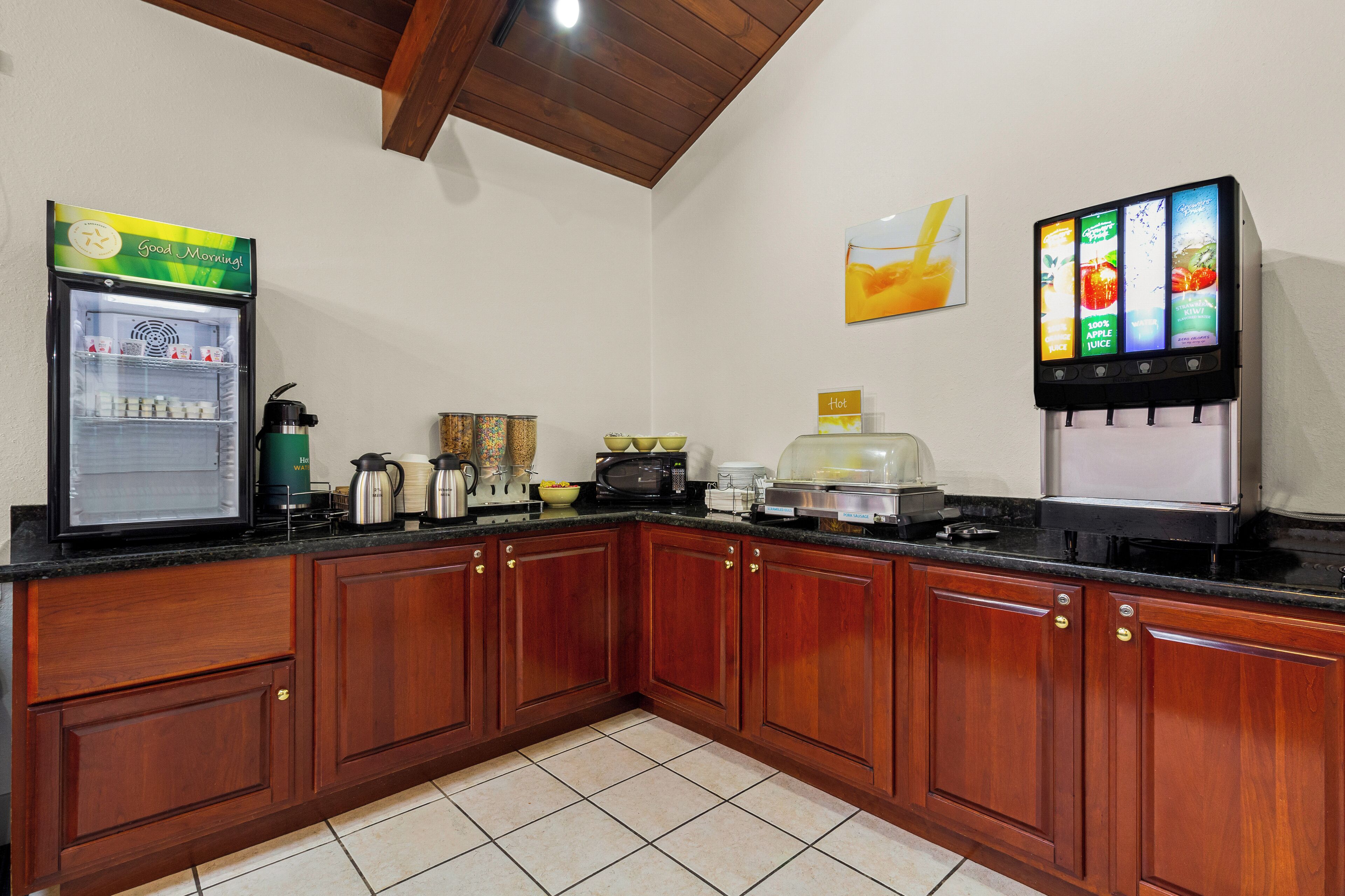 A kitchen with wooden cabinets, a refrigerator, a microwave, a coffee maker, and a juice dispenser.