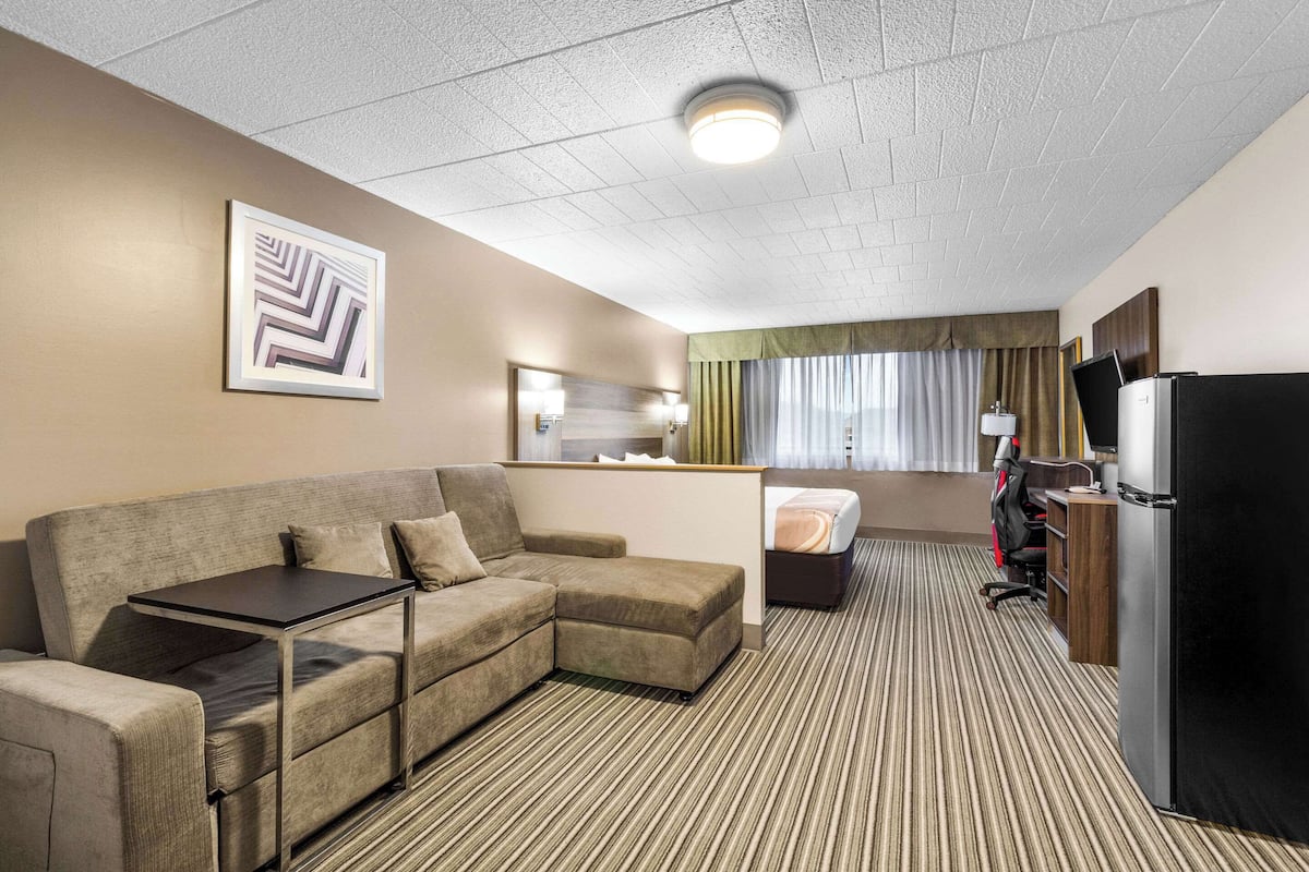 Ett hotellrum med en soffa, ett litet bord, ett kylskåp, ett skrivbord och ett fönster med gardiner.