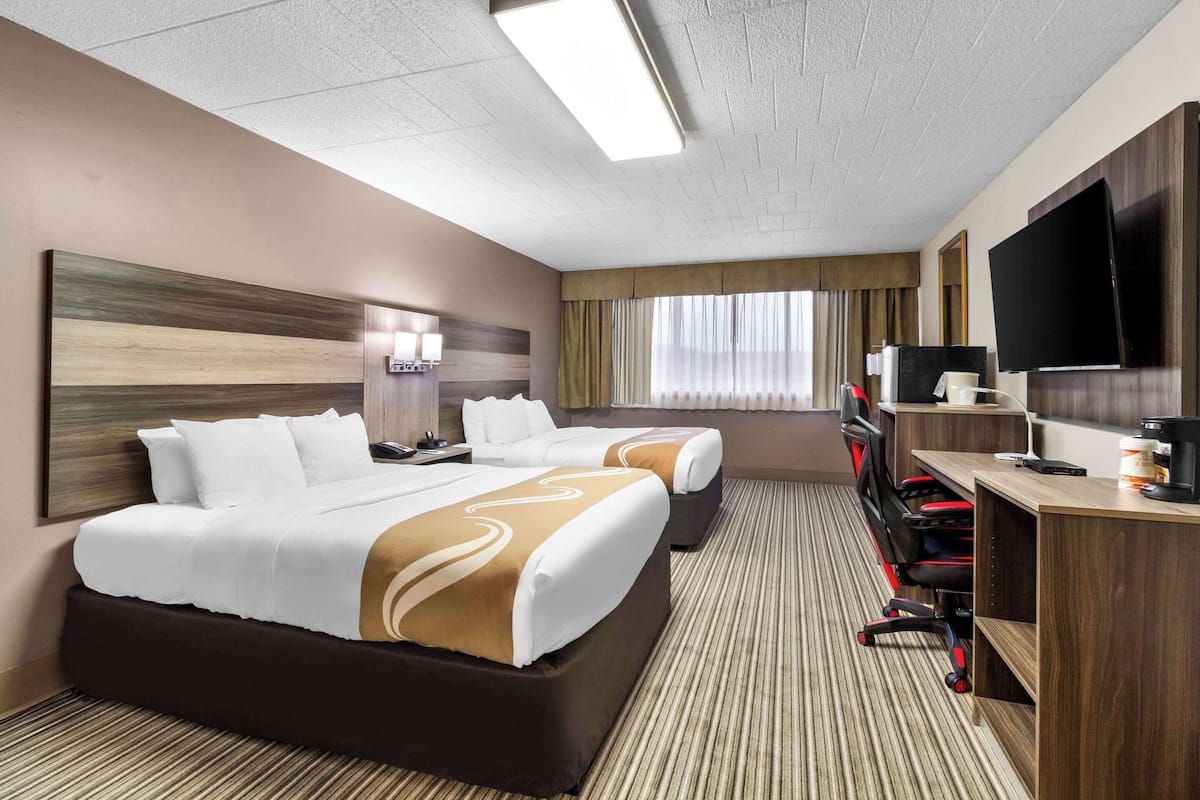 Ett hotellrum med två sängar, ett skrivbord med en dator, en tv och ett stort fönster med gardiner.