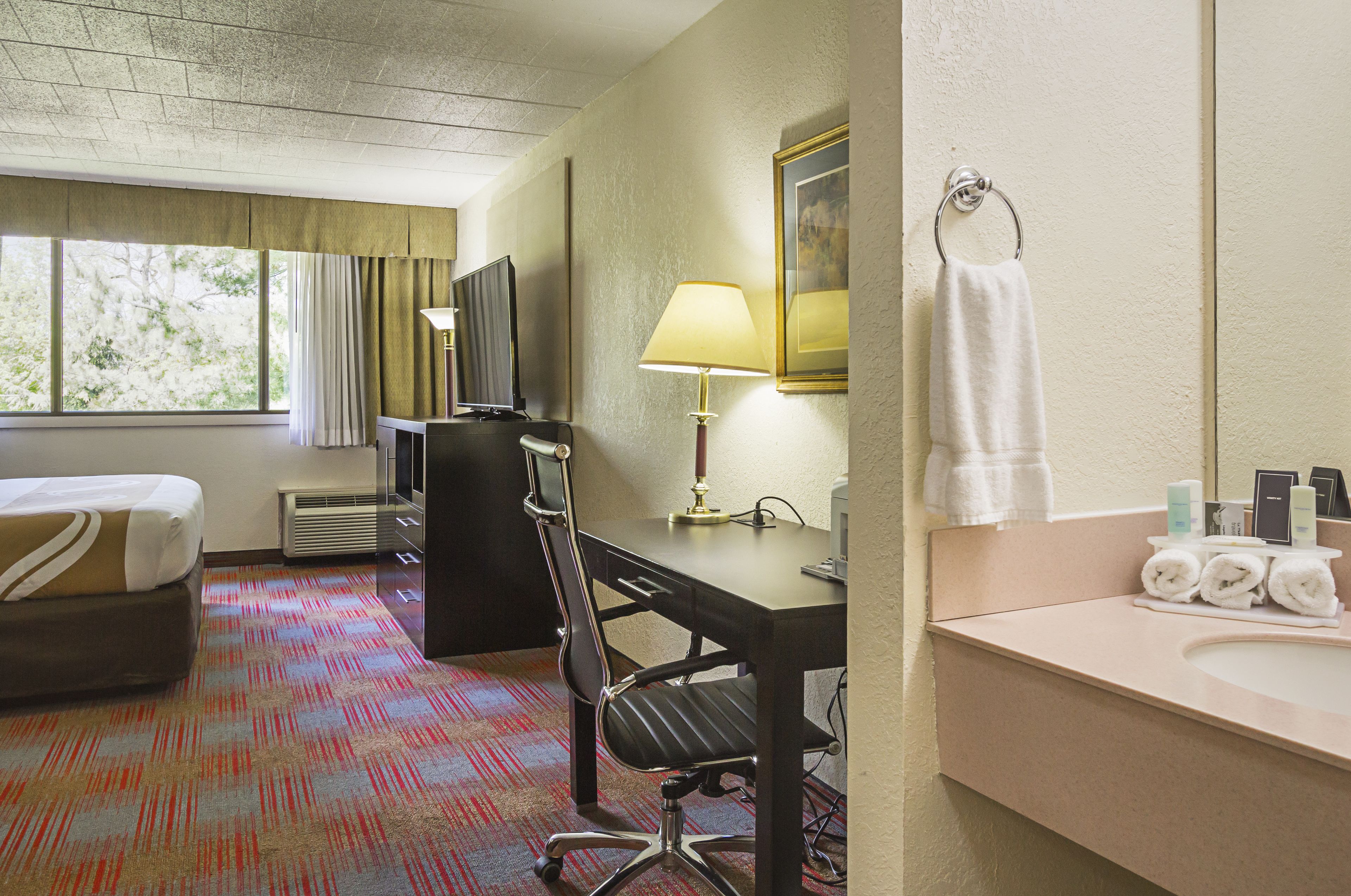 A hotel room with a bed, desk, chair, lamp, and a bathroom with towels and toiletries.