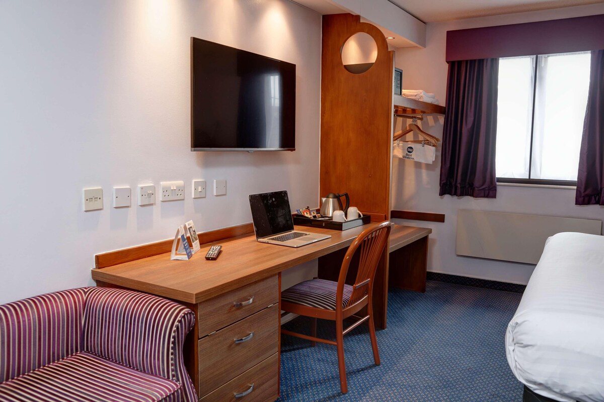 A hotel room with a bed, a desk with a laptop, a chair, a TV, and a window with curtains.