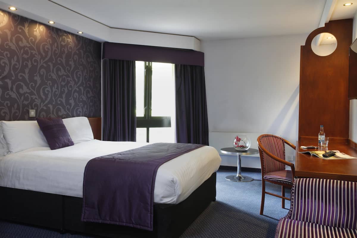 A hotel room with a large bed, a small table with a chair, and a window with curtains.