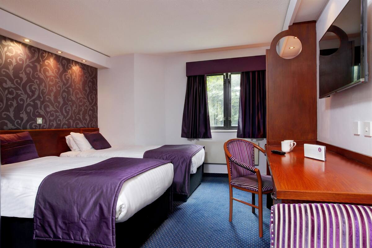 A hotel room with two beds, a wooden desk, a chair, a television, and a window with curtains.