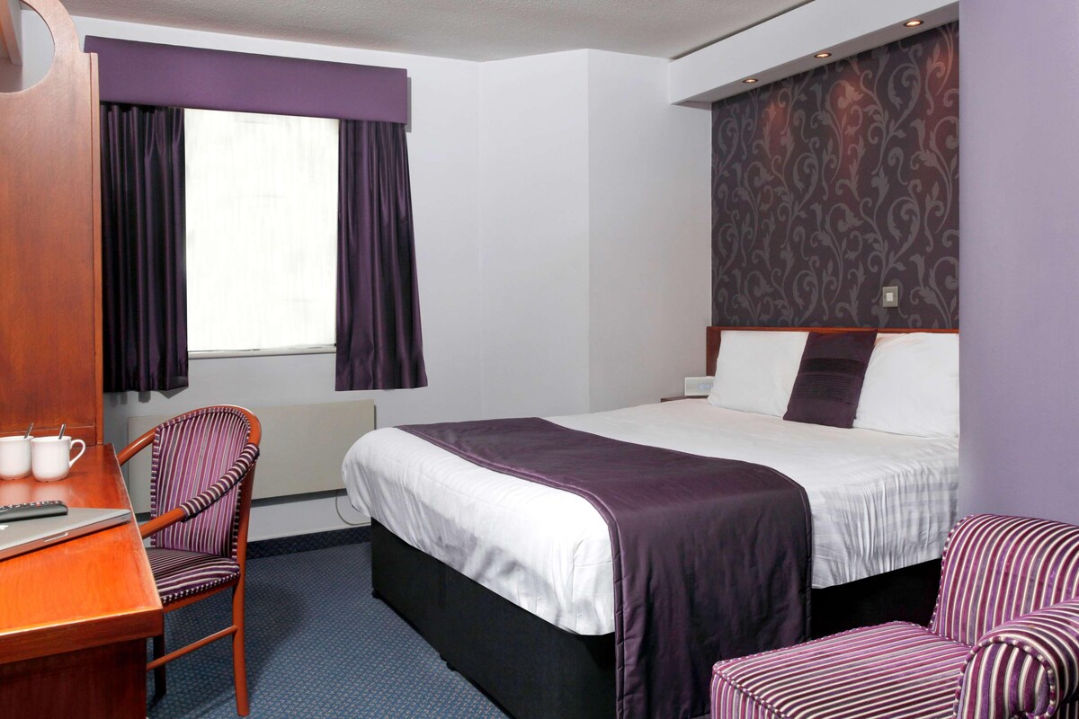 A hotel room with a bed, a desk with a chair, and a window with curtains.