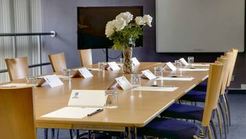 A conference room with a long table, chairs, a vase with white flowers, nameplates, and a projector screen.