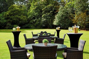 An outdoor seating area with round tables and wicker chairs, surrounded by a well-maintained lawn and trees.