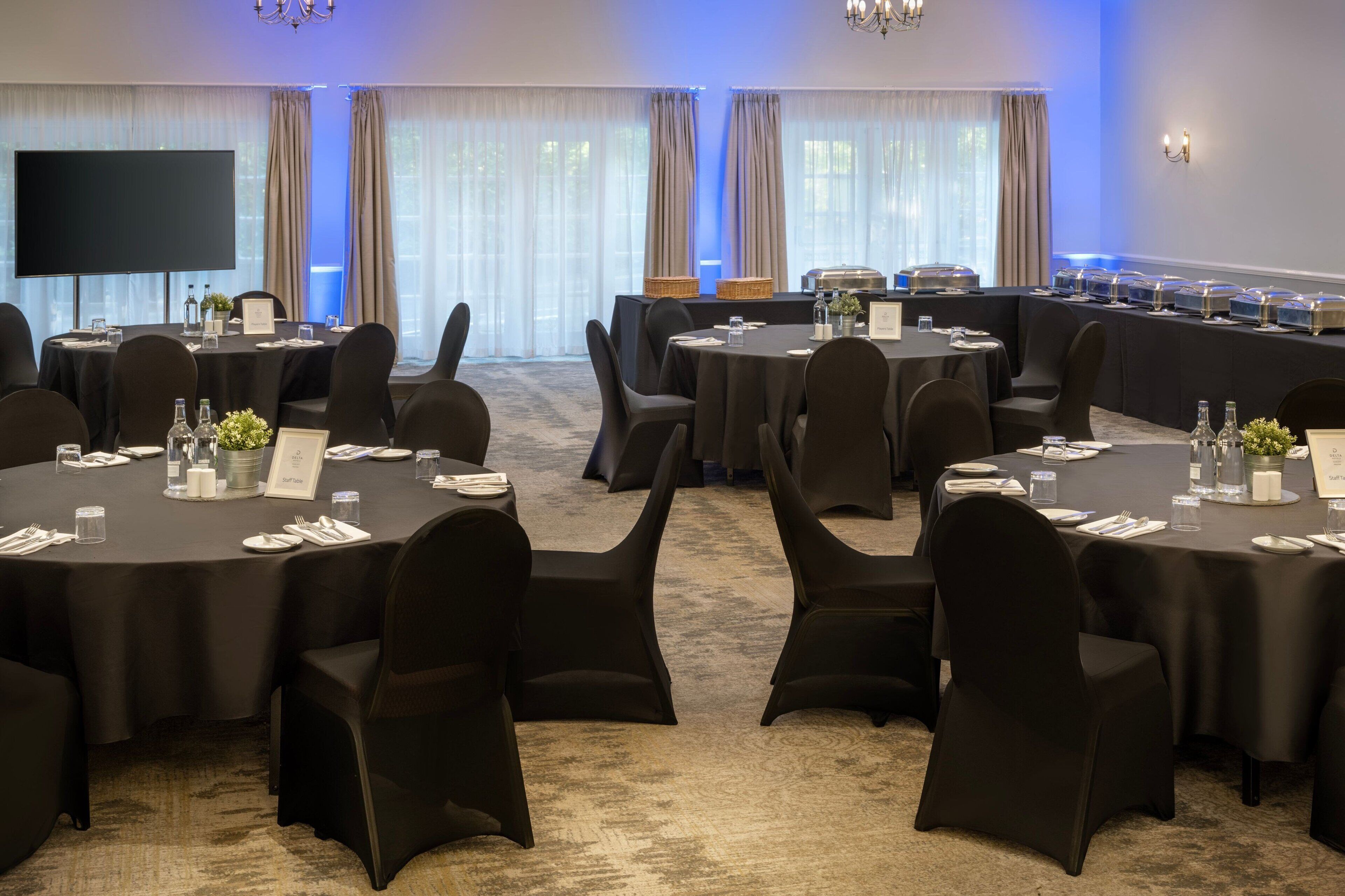 A conference room with multiple round tables, black chairs, a large flat-screen TV, and a buffet setup.