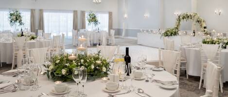 Un salón de banquetes con mesas redondas dispuestas para un evento formal, con manteles blancos, centros de mesa florales e iluminación con velas.