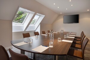 Una sala de conferencias con una larga mesa, sillas, un televisor de pantalla plana y ventanas amplias que dan a zonas verdes.