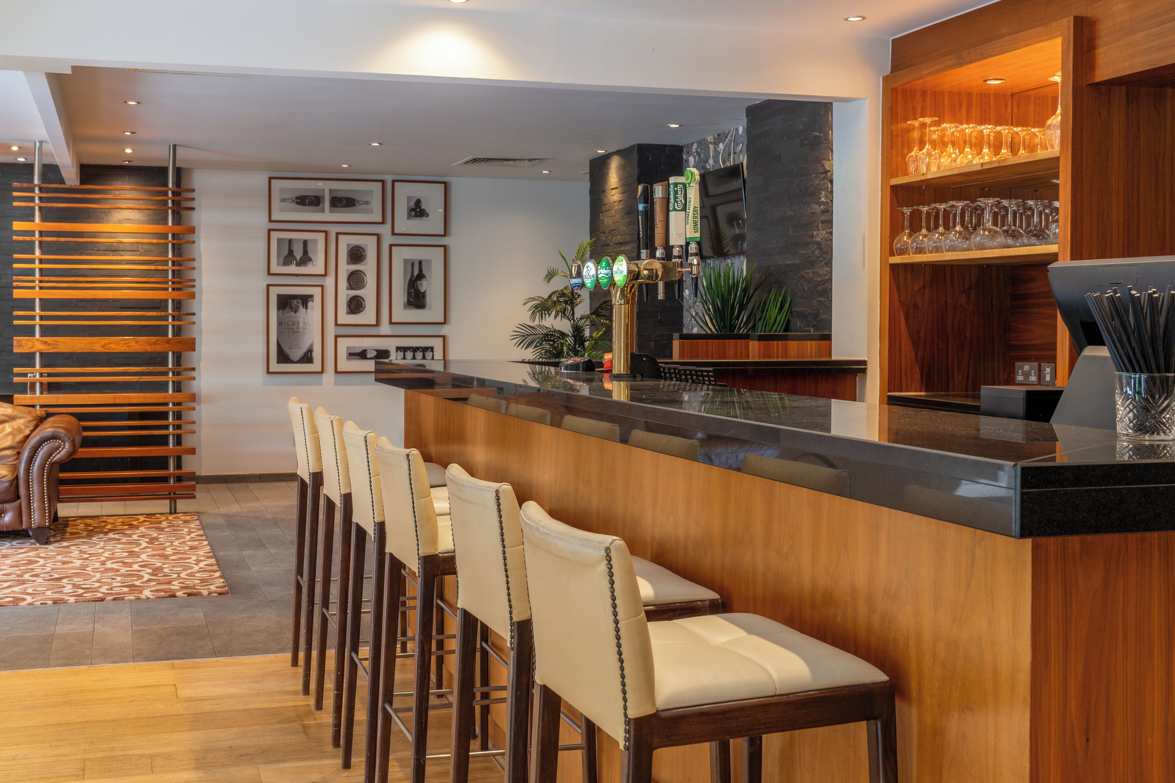 Un espace bar avec un comptoir en bois, des tabourets de bar et une étagère où sont rangés les verres.