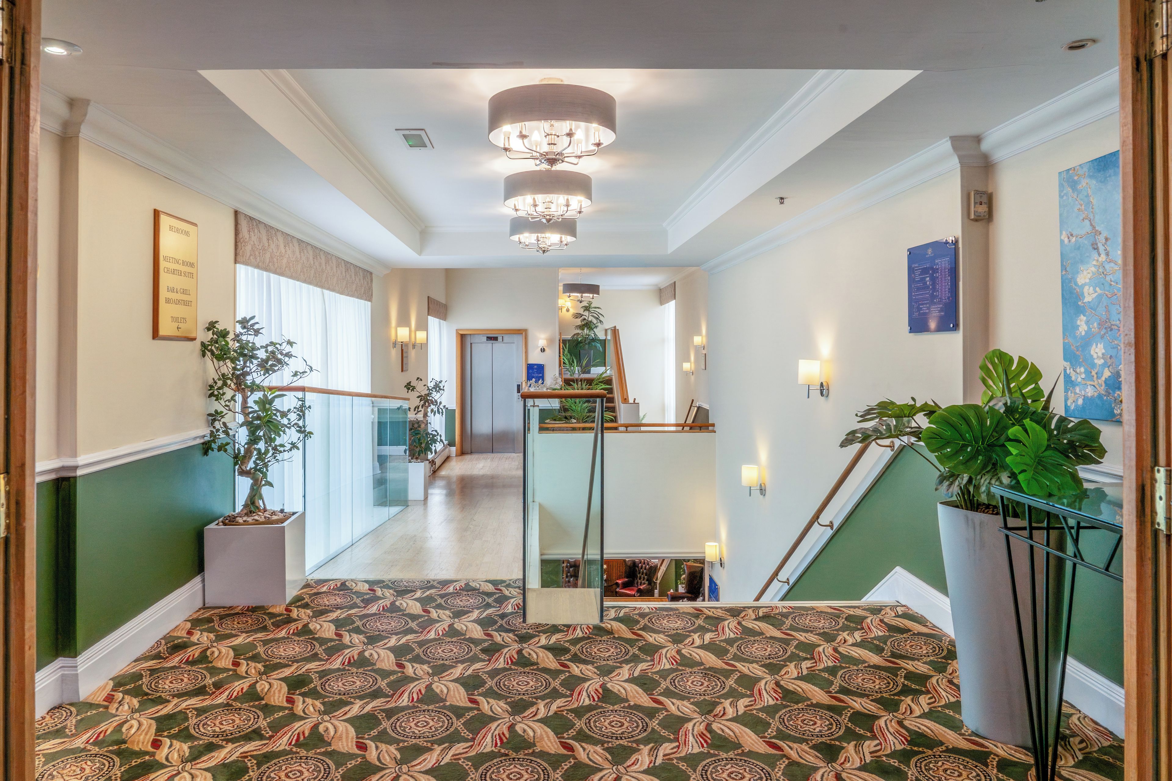 Un pasillo de hotel con alfombra estampada, un candelabro, una escalera y una planta en maceta.