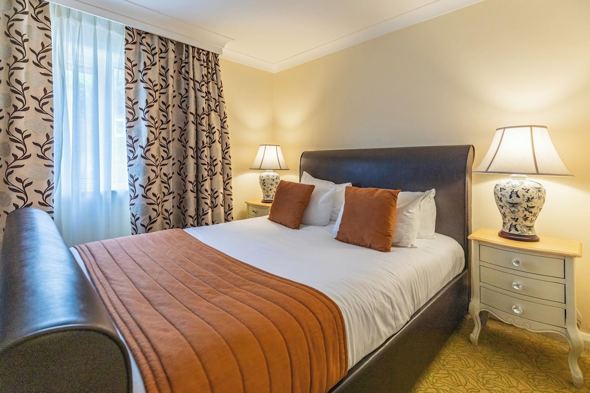 A neatly made bed with a brown blanket, two bedside tables with lamps, and a window with patterned curtains.