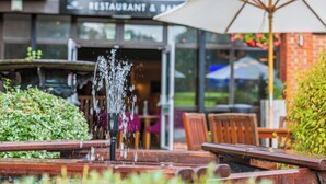 An outdoor seating area with a water feature, wooden tables, and chairs. In the background, there is a restaurant with a sign that reads "RESTAURANT & BAR". - Meon Valley Hotel, Spa & Golf (Southampton)