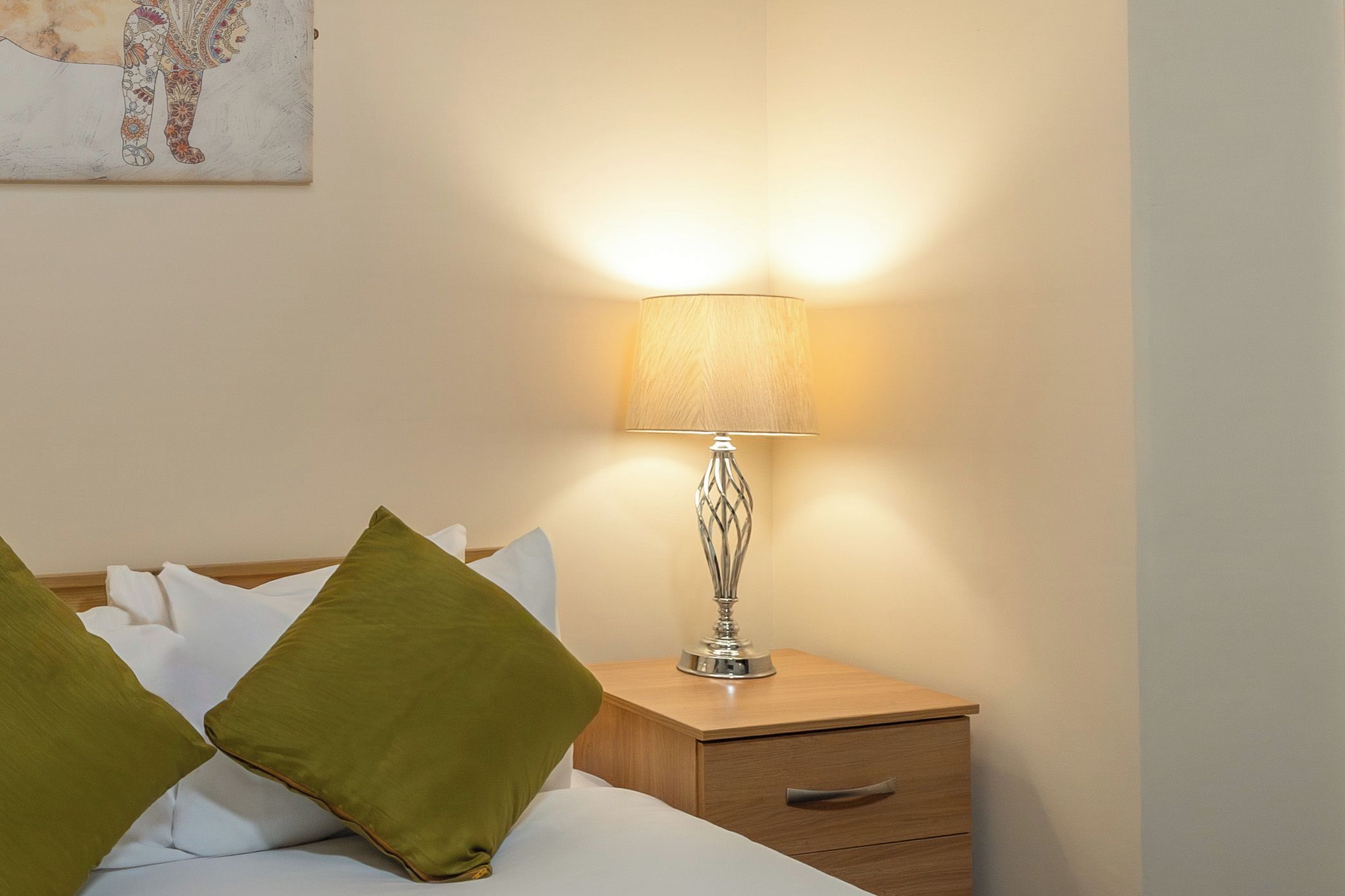 Ein ordentlich bezogenes Bett mit grünen Kissen, ein Nachttisch mit Lampe und ein Gemälde an der Wand.