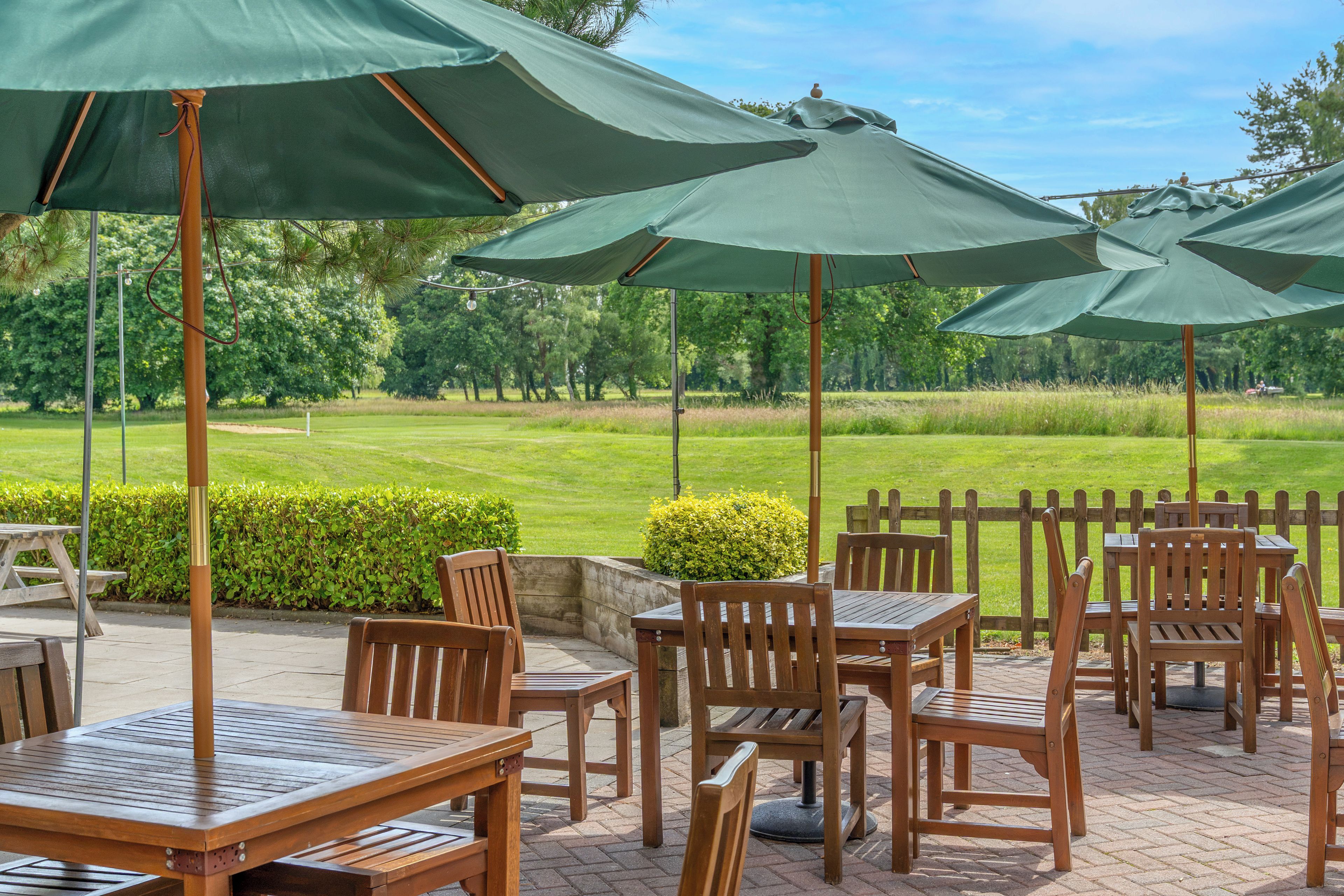 Un patio al aire libre con mesas y sillas de madera, sombrillas verdes y vistas a un campo de golf.