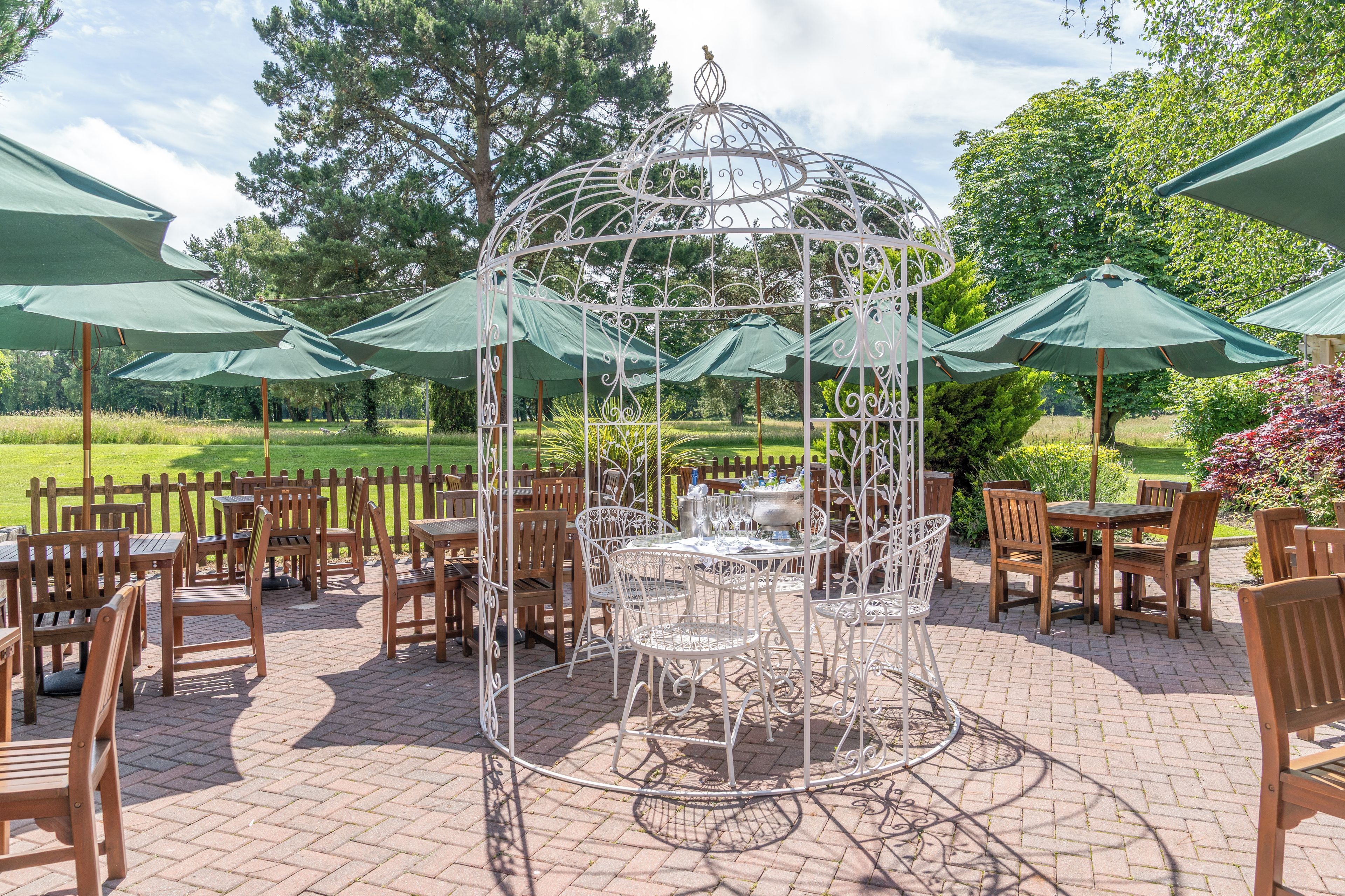 A gazebo with a metal frame and a table in the center, surrounded by wooden chairs and tables with green umbrellas.