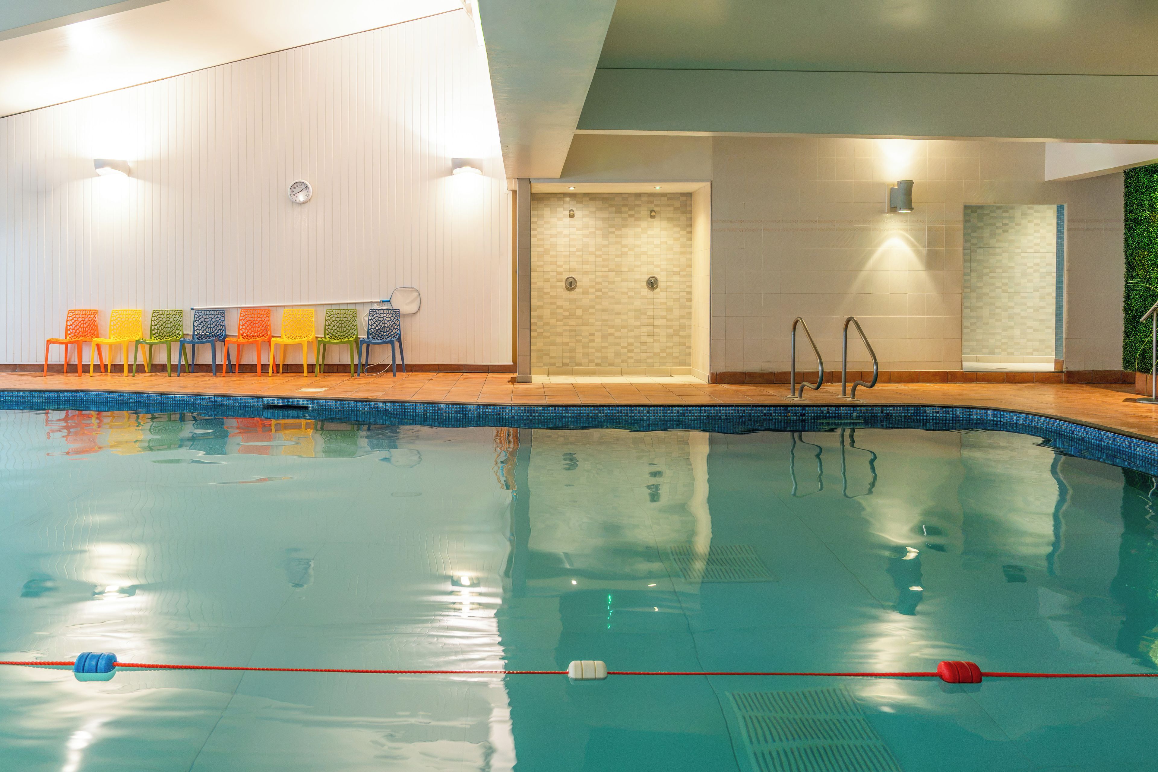 An indoor swimming pool with a tiled edge, a changing room with a mirror, and a row of colorful chairs.