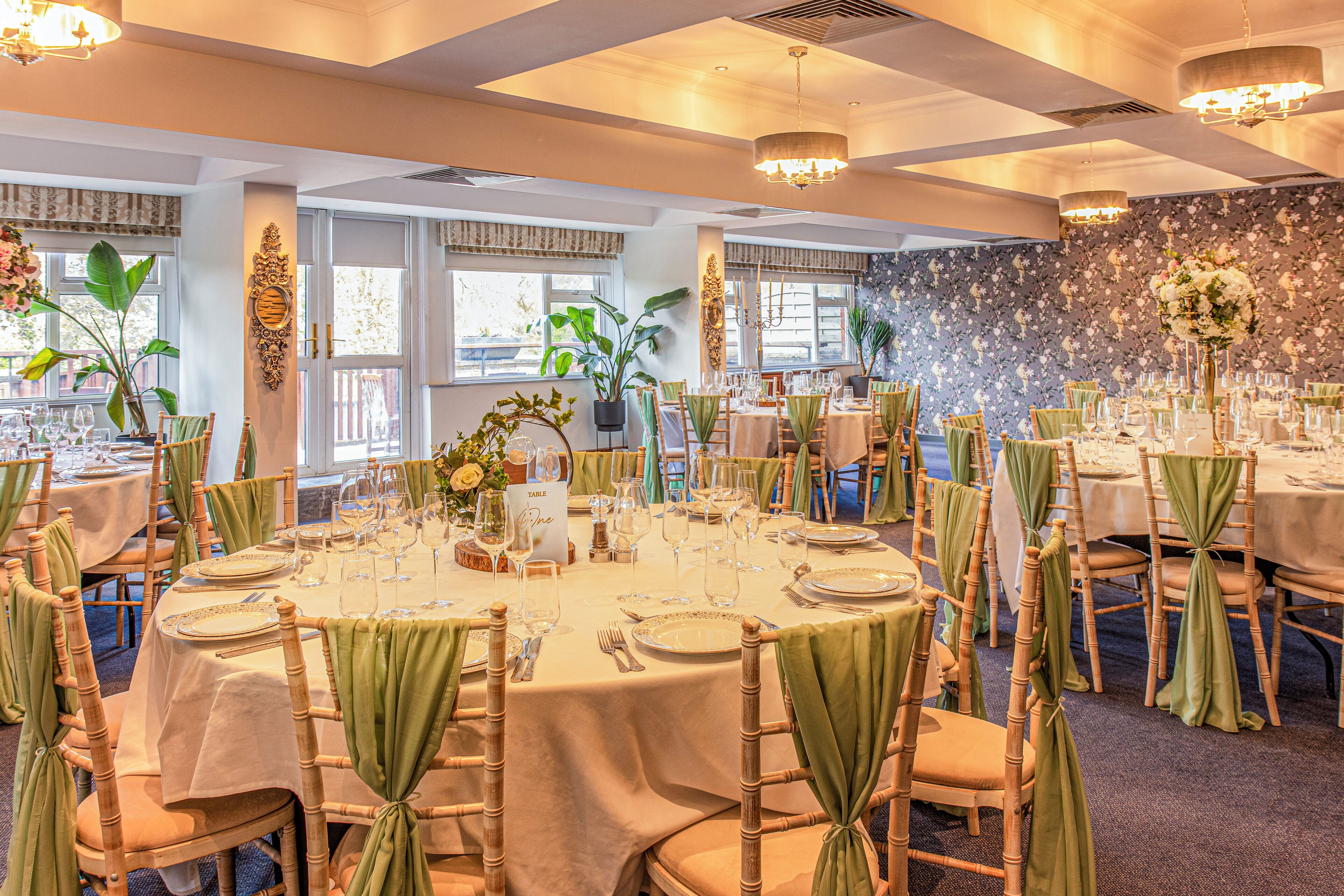 Un salón de banquetes con mesas redondas, fundas de sillas verdes y centros de mesa florales.