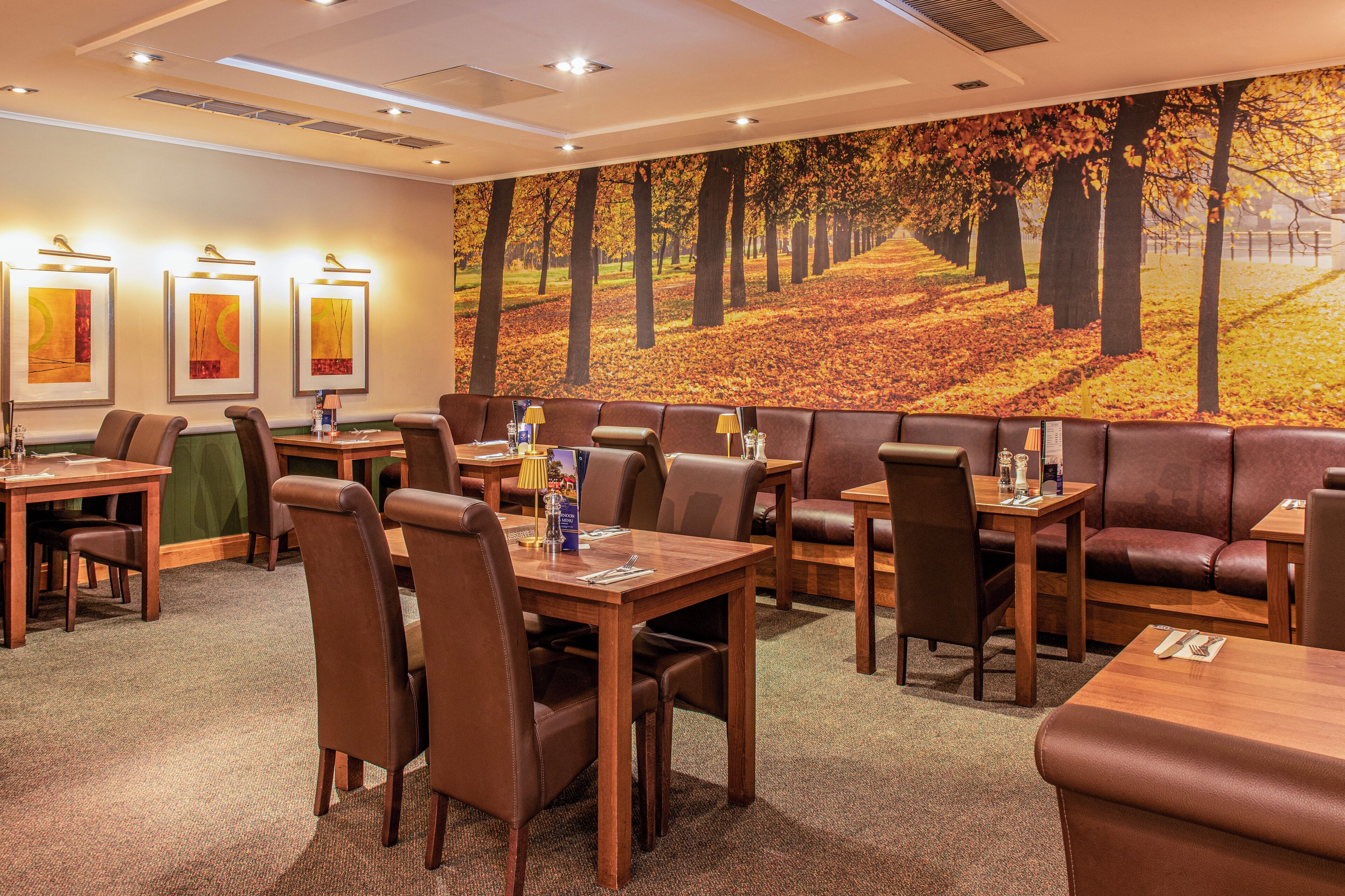 Un comedor de hotel con varias mesas y cabinas, suelo alfombrado y un gran mural con motivos otoñales en la pared.