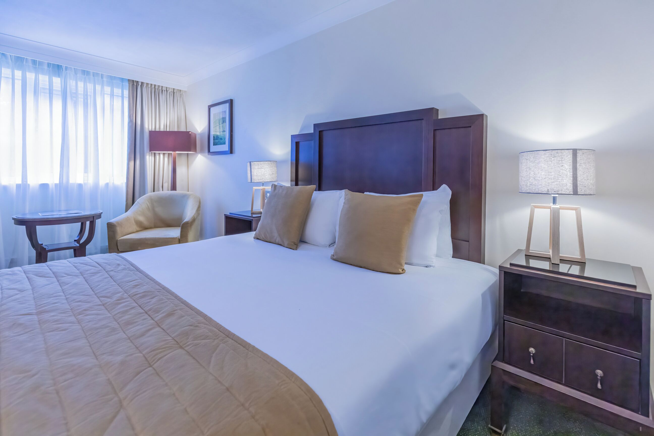 A hotel room with a large bed, a chair, a nightstand with a lamp, and a window with curtains.