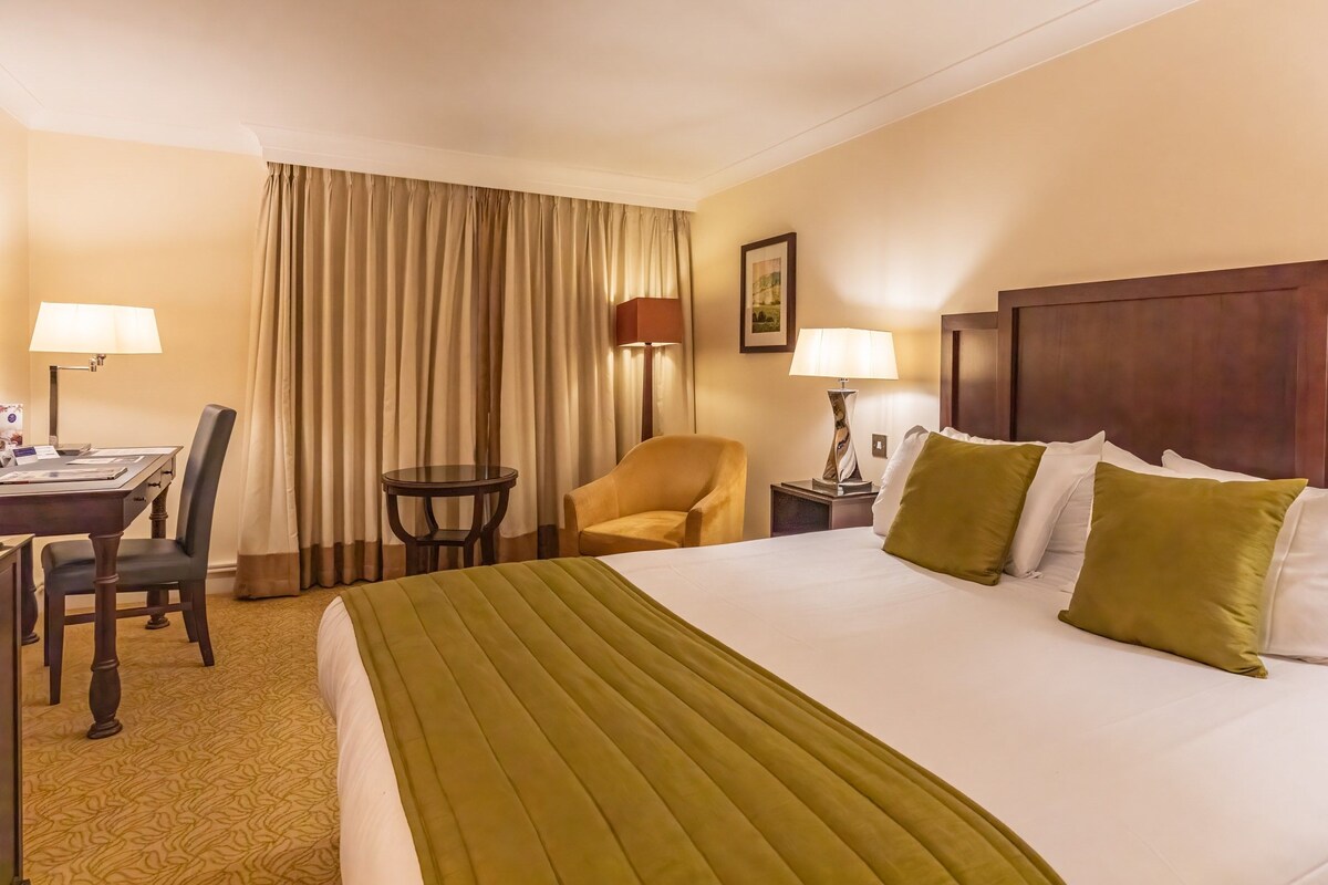 A hotel room with a large bed, two bedside lamps, a desk with a chair, a small round table, and a large window with curtains.