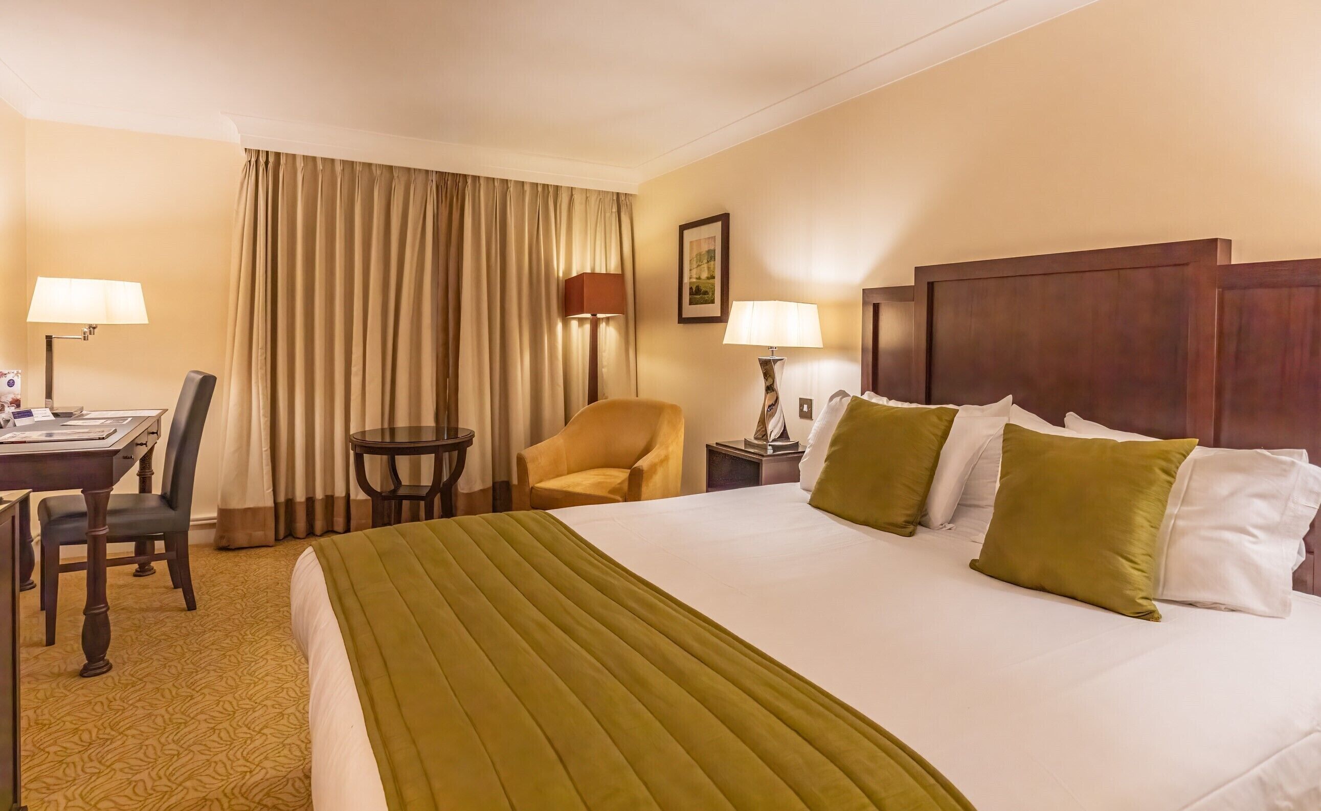 Una camera d'albergo con un letto grande, due abat-jour, una scrivania con una sedia, un piccolo tavolino rotondo e un'ampia finestra con tende.