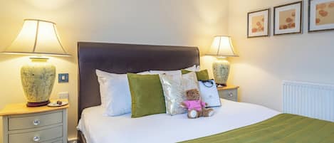 Una cama bien hecha con un edredón verde, dos lámparas de noche, un oso de peluche y un jarrón decorativo.