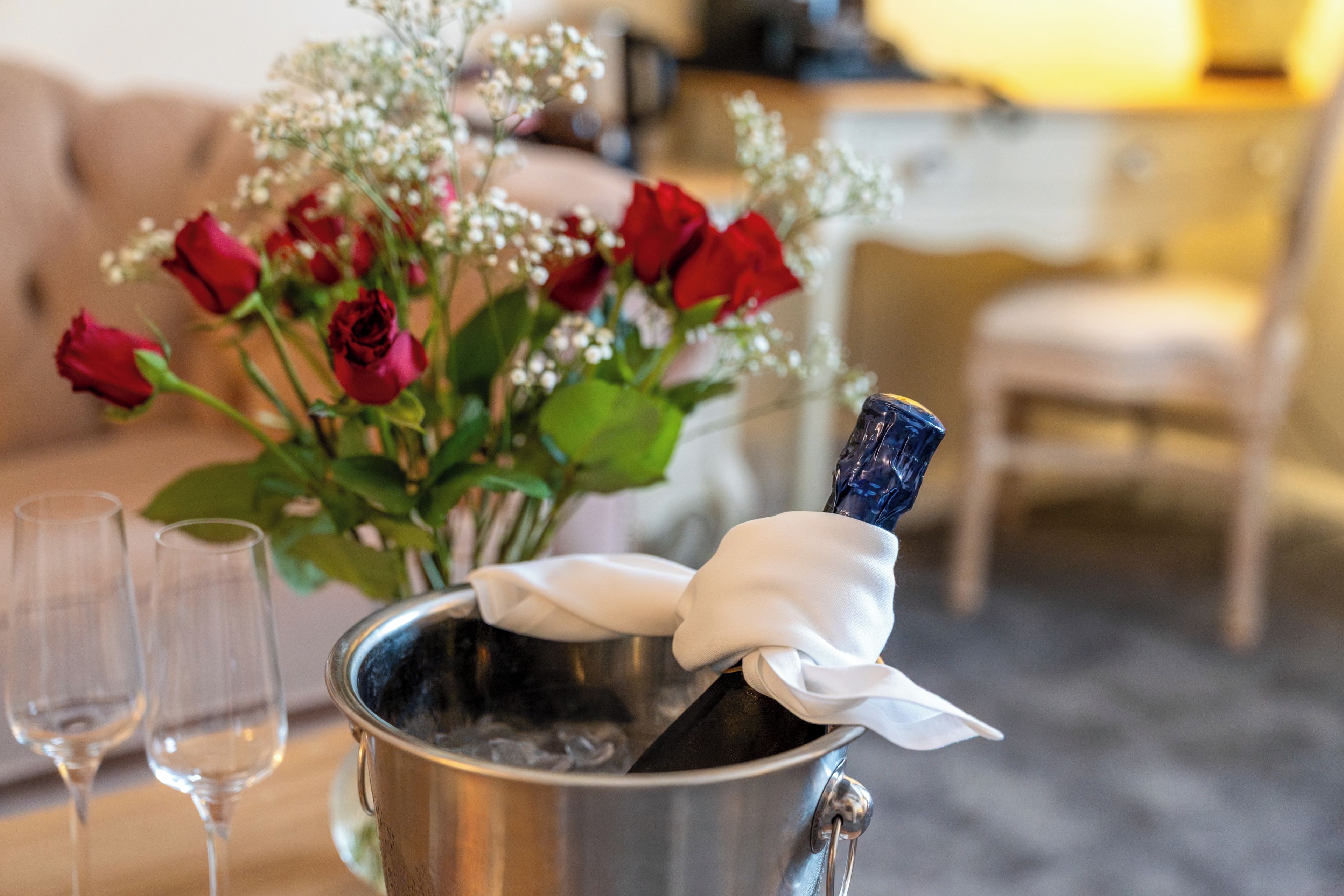 A champagne bottle in a metal bucket with a white cloth, red roses, and two empty glasses on a table.