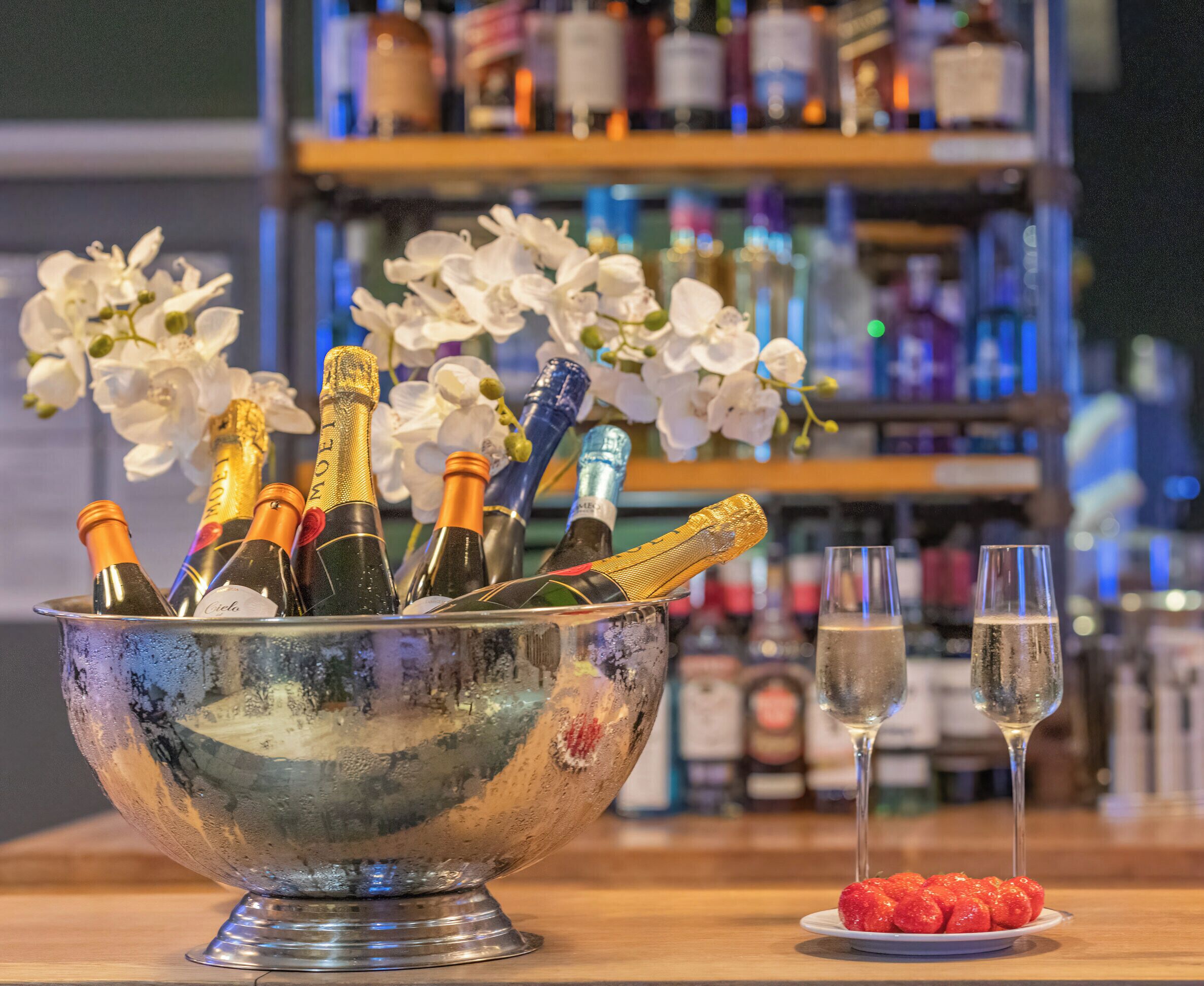 A bar area with a metal bucket containing champagne bottles, two glasses of champagne, and a plate of strawberries.