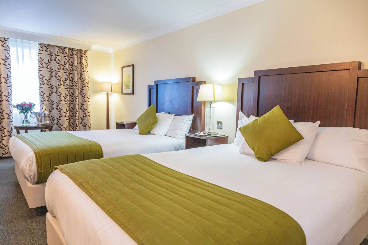 A hotel room with two beds, each with a green blanket, white bedding, and a wooden headboard. There are bedside tables with lamps and a small table with a vase of flowers.