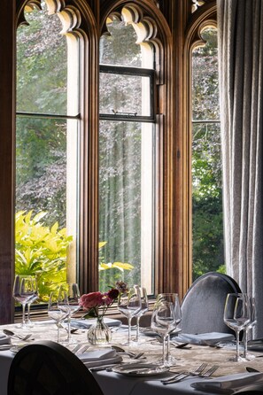 Une table à manger dressée avec des verres à vin, des assiettes et des couverts, agrémentée d’un vase de fleurs et offrant une vue sur la verdure à travers de grandes fenêtres.