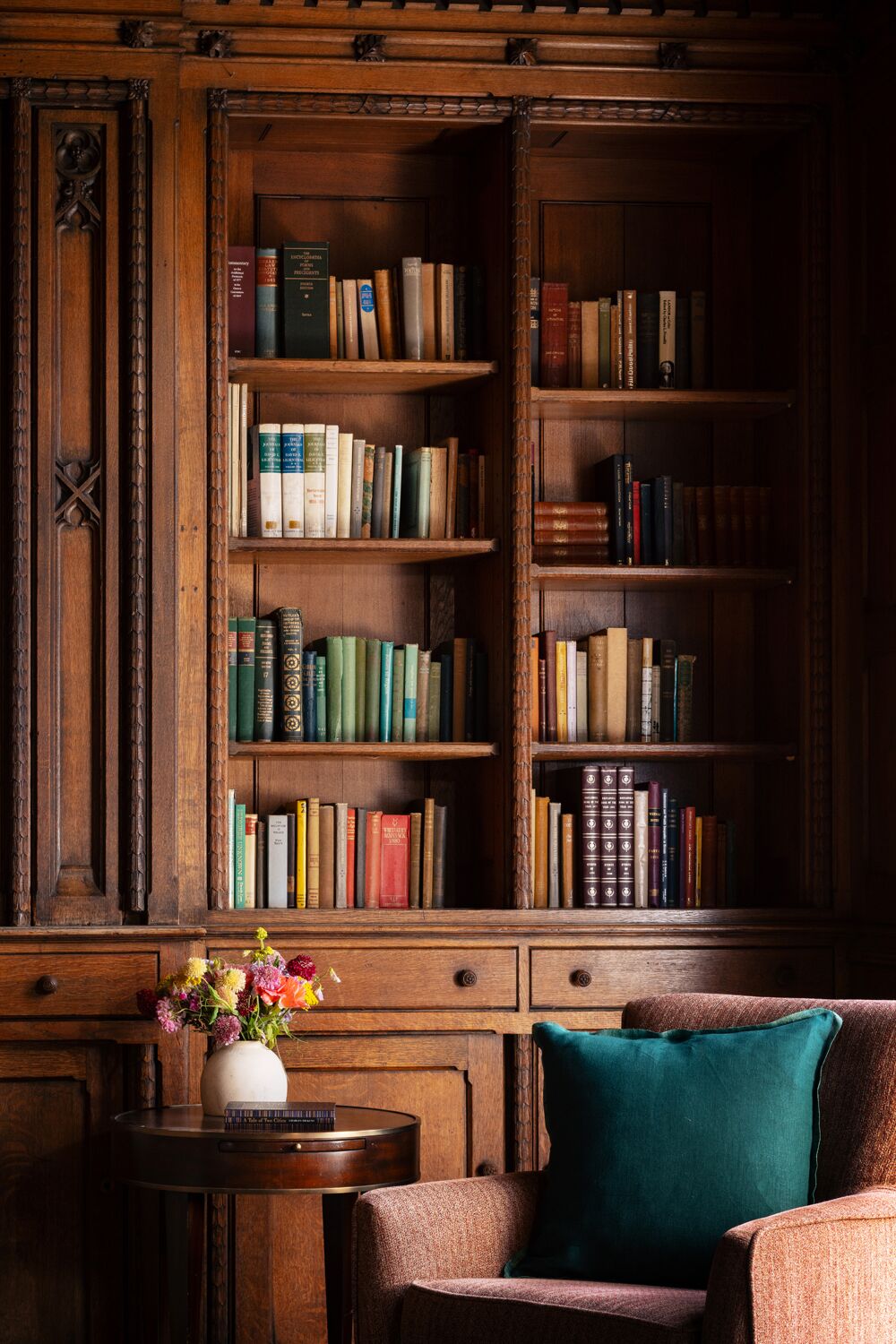Una estantería de madera llena de libros, una pequeña mesa redonda con un jarrón de flores y una silla acolchada con un cojín azul turquesa.