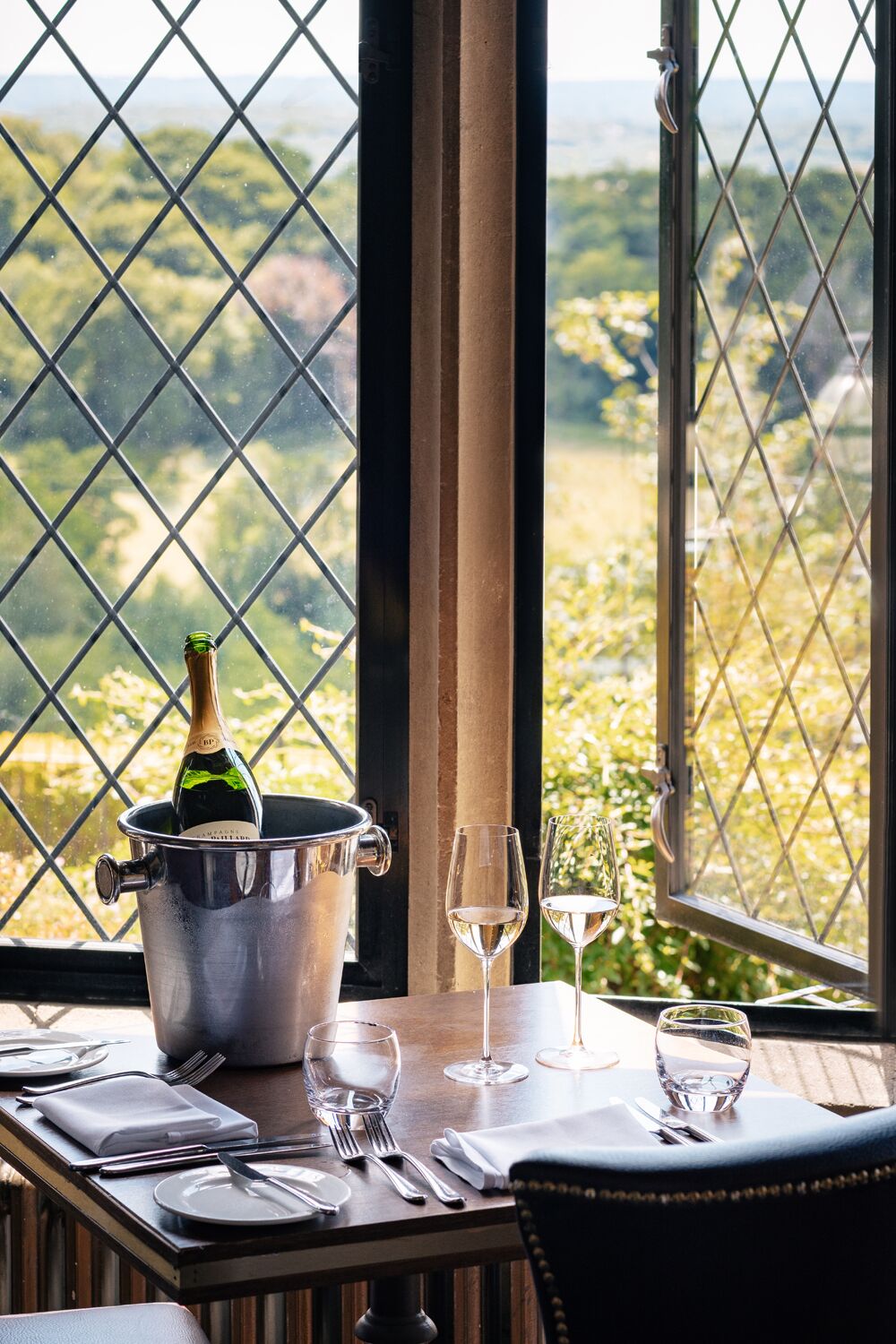 A dining table set with a bottle of champagne, two wine glasses, and a view of a vineyard through a diamond-patterned window.