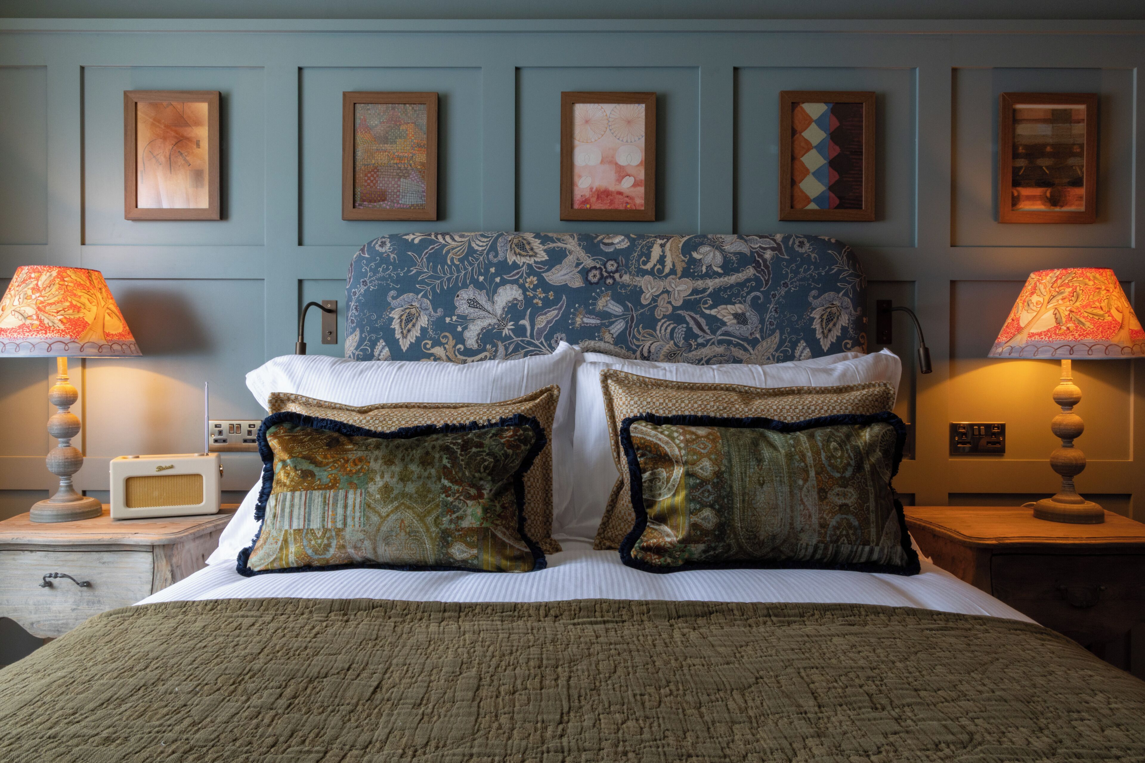 Una cama bien hecha con almohadones estampados y dos lámparas de noche, un reloj de pared y cuadros en la pared.