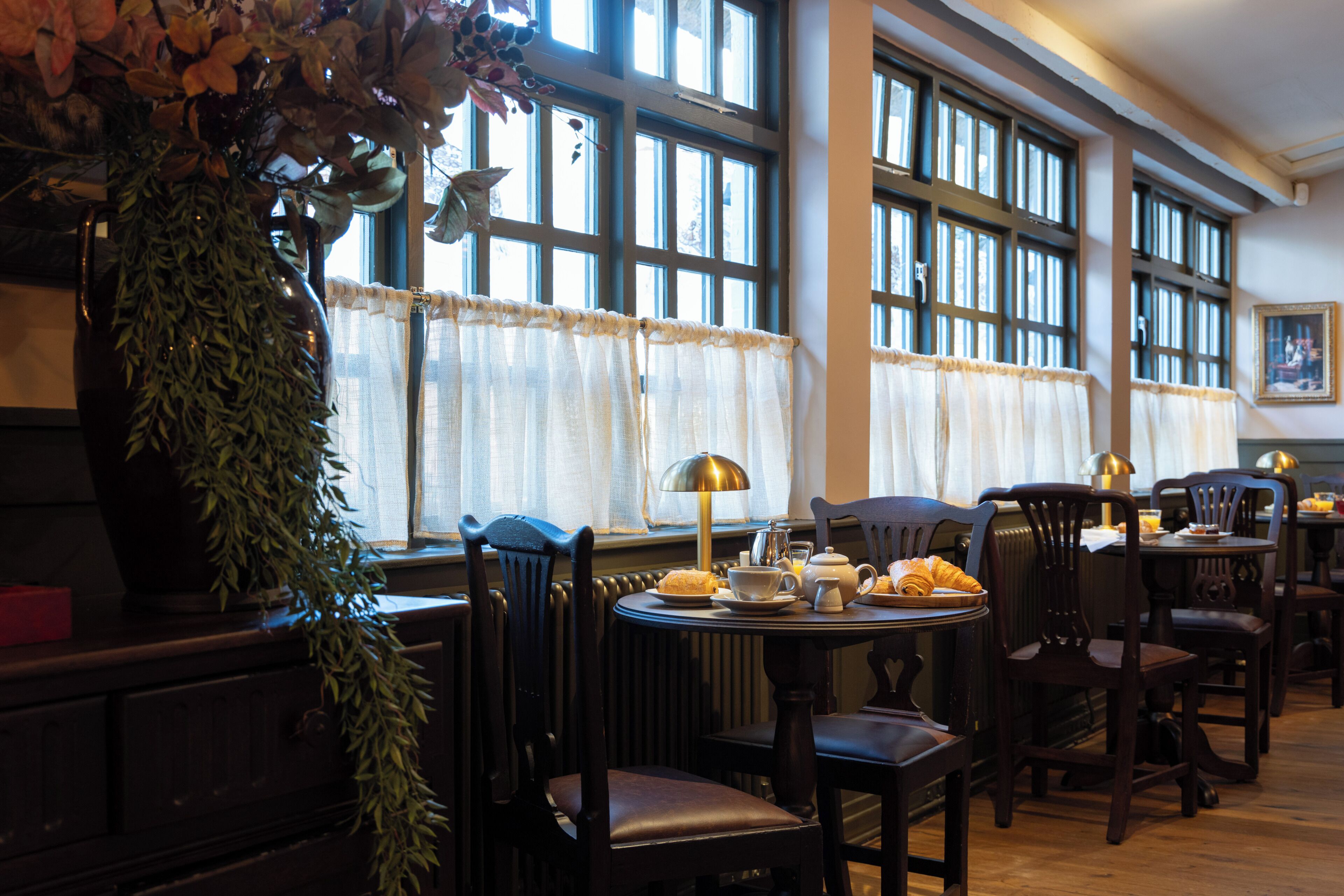 A dining area with a table set for two, chairs, a potted plant, and a window with curtains.