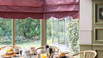 A dining area with a round table, wicker chairs, and a view of greenery through large windows.