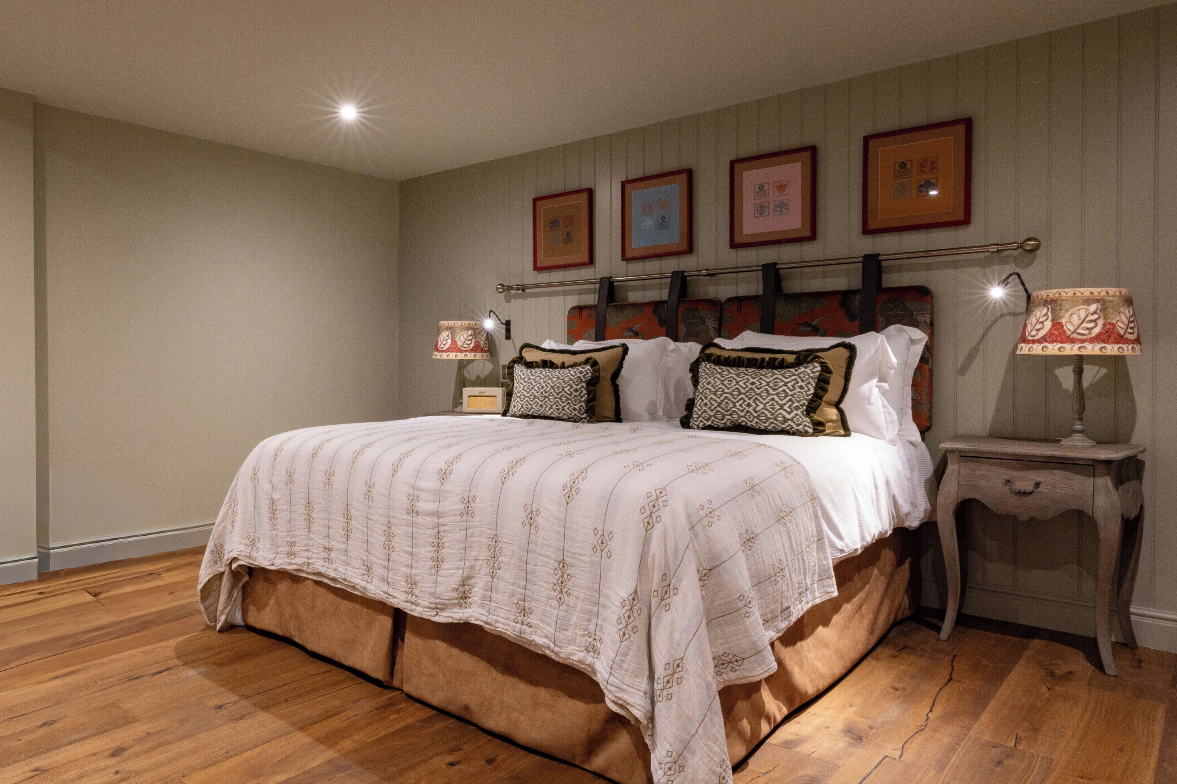 Un dormitorio con una cama grande, suelo de madera, cabecera con obra de arte, lámparas de noche y cuadros en la pared.