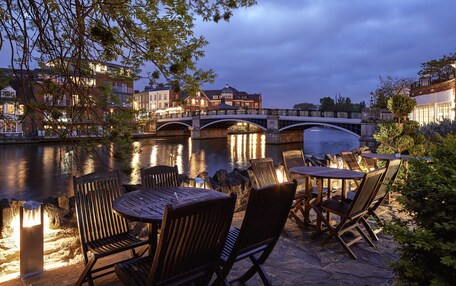 An outdoor seating area with tables and chairs by a river, a bridge, and buildings in the background.