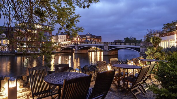 An outdoor seating area with tables and chairs by a river, a bridge, and buildings in the background. - Sir Christopher Wren Hotel (Windsor)