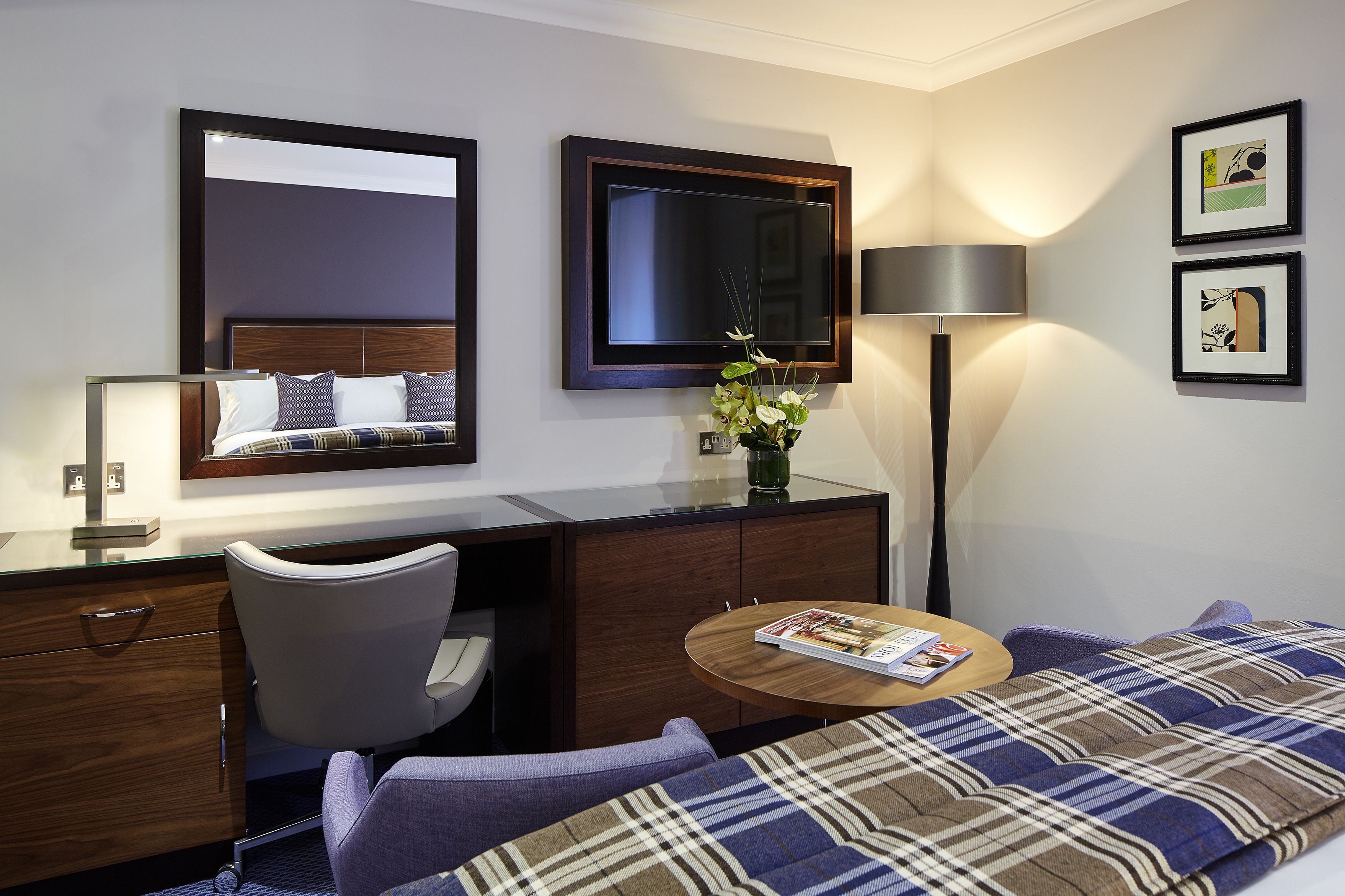 Una habitación de hotel con cama, escritorio, silla, lámpara y dos cuadros en la pared.