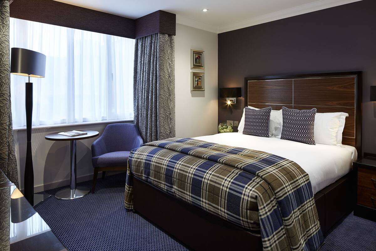 A hotel room with a large bed, a bedside table, a chair, a floor lamp, and a window with curtains.