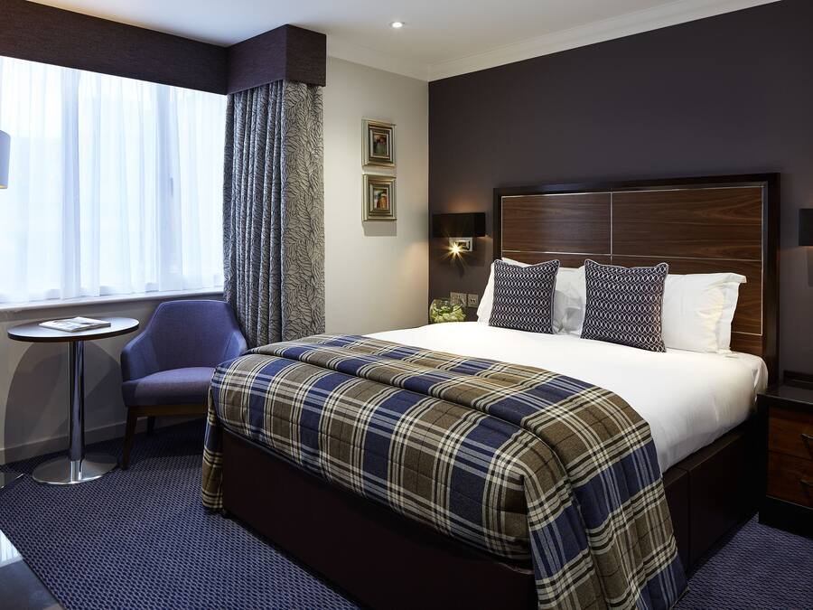 A hotel room with a large bed, a bedside table, a chair, a floor lamp, and a window with curtains.