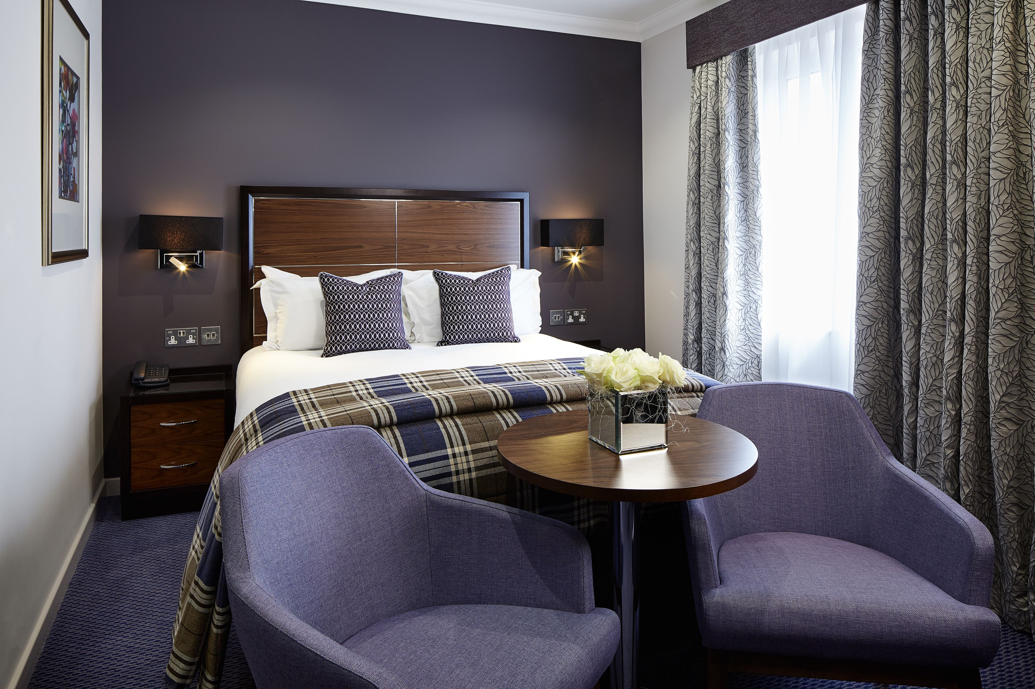 A hotel room with a large bed, two bedside tables, a small table with chairs, and a window with curtains.