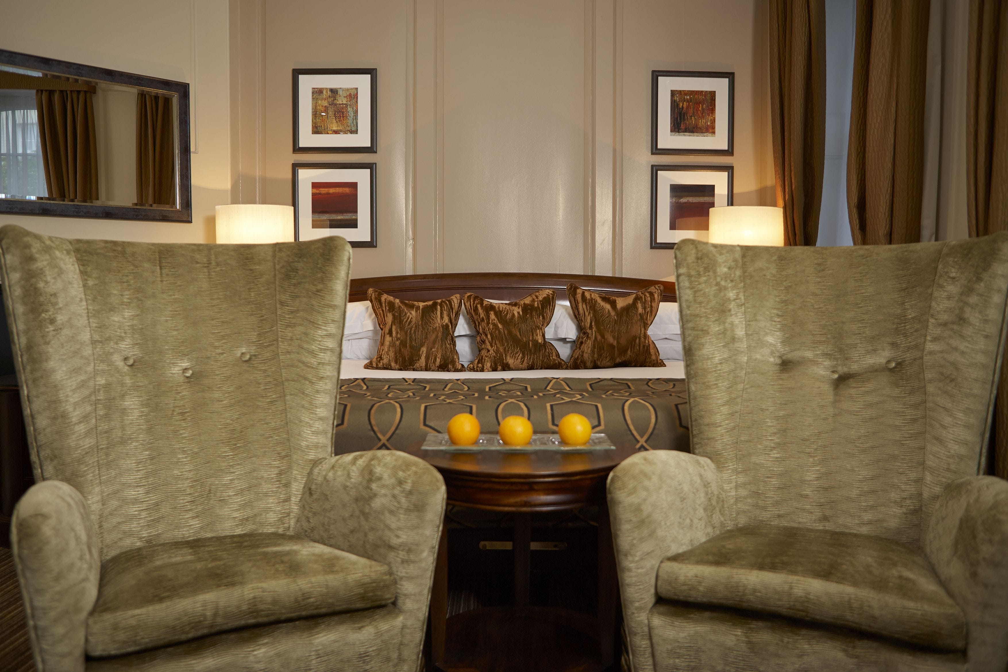 A hotel room with a bed, two armchairs, a small table with three lemons, and framed pictures on the wall.