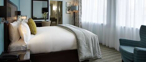 A hotel room with a large bed, a desk with a telephone, a chair, a mirror, and a window with sheer curtains.