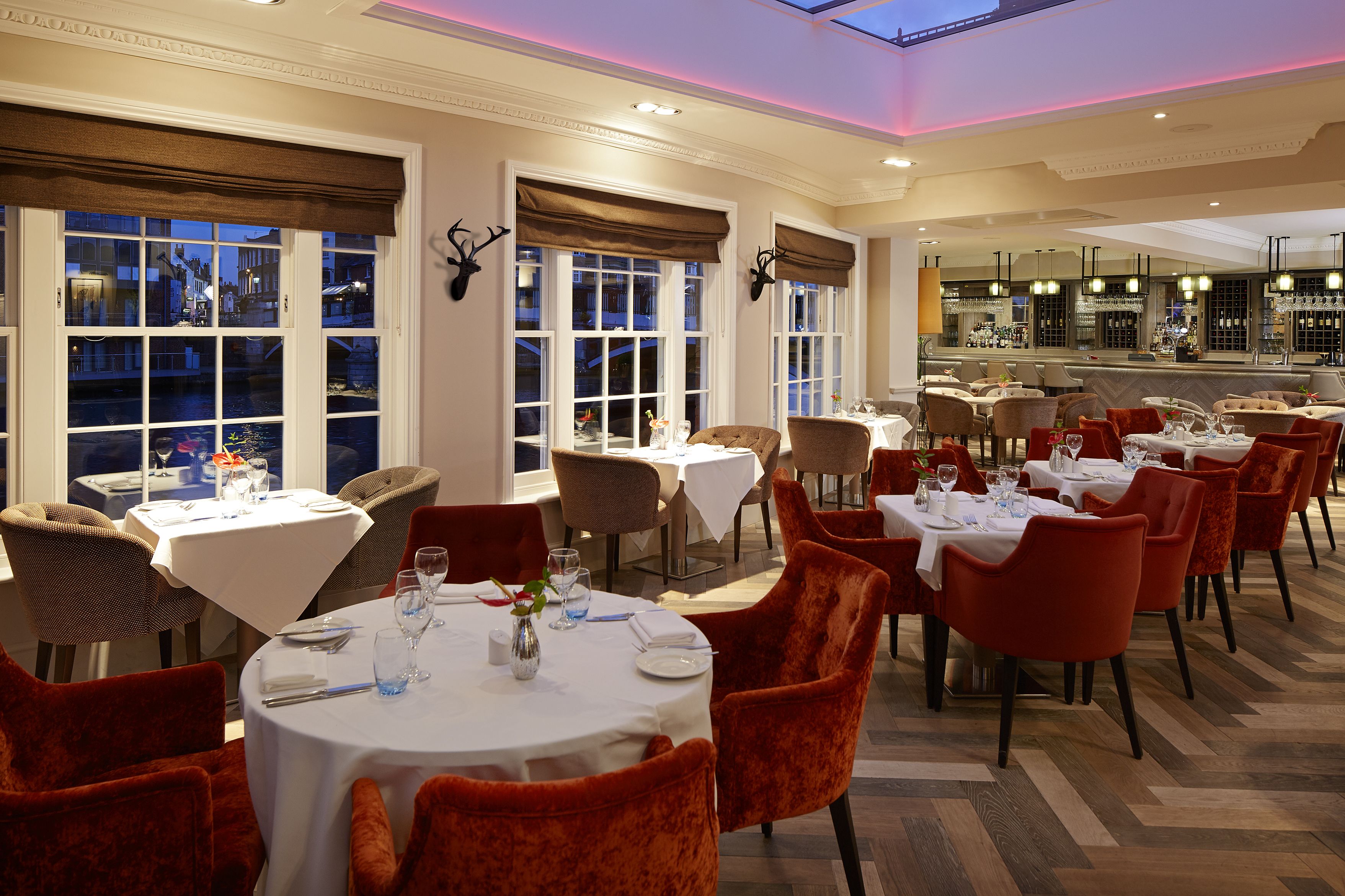 A restaurant with multiple round tables, red velvet chairs, and a bar area.