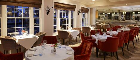 A restaurant with multiple round tables, red velvet chairs, and a bar area.