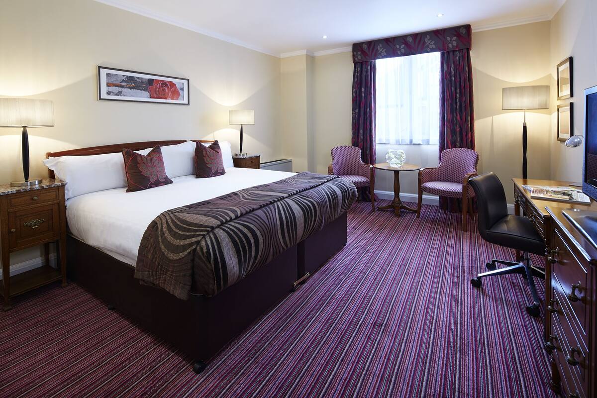 A hotel room with a large bed, two chairs, a desk, and a television.